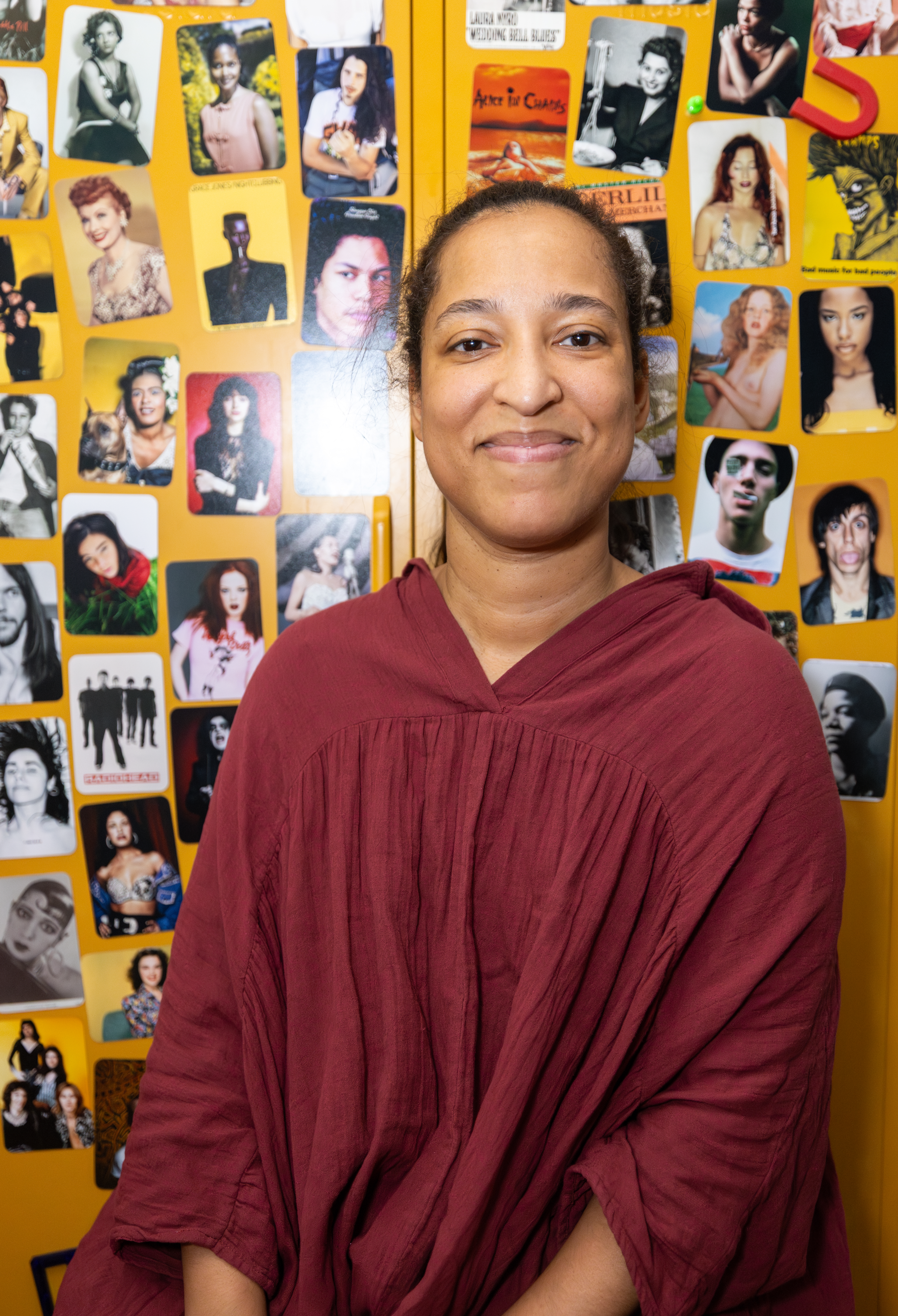 woman sits and smiles in front of yellow locker with small photos of celebrities and artists