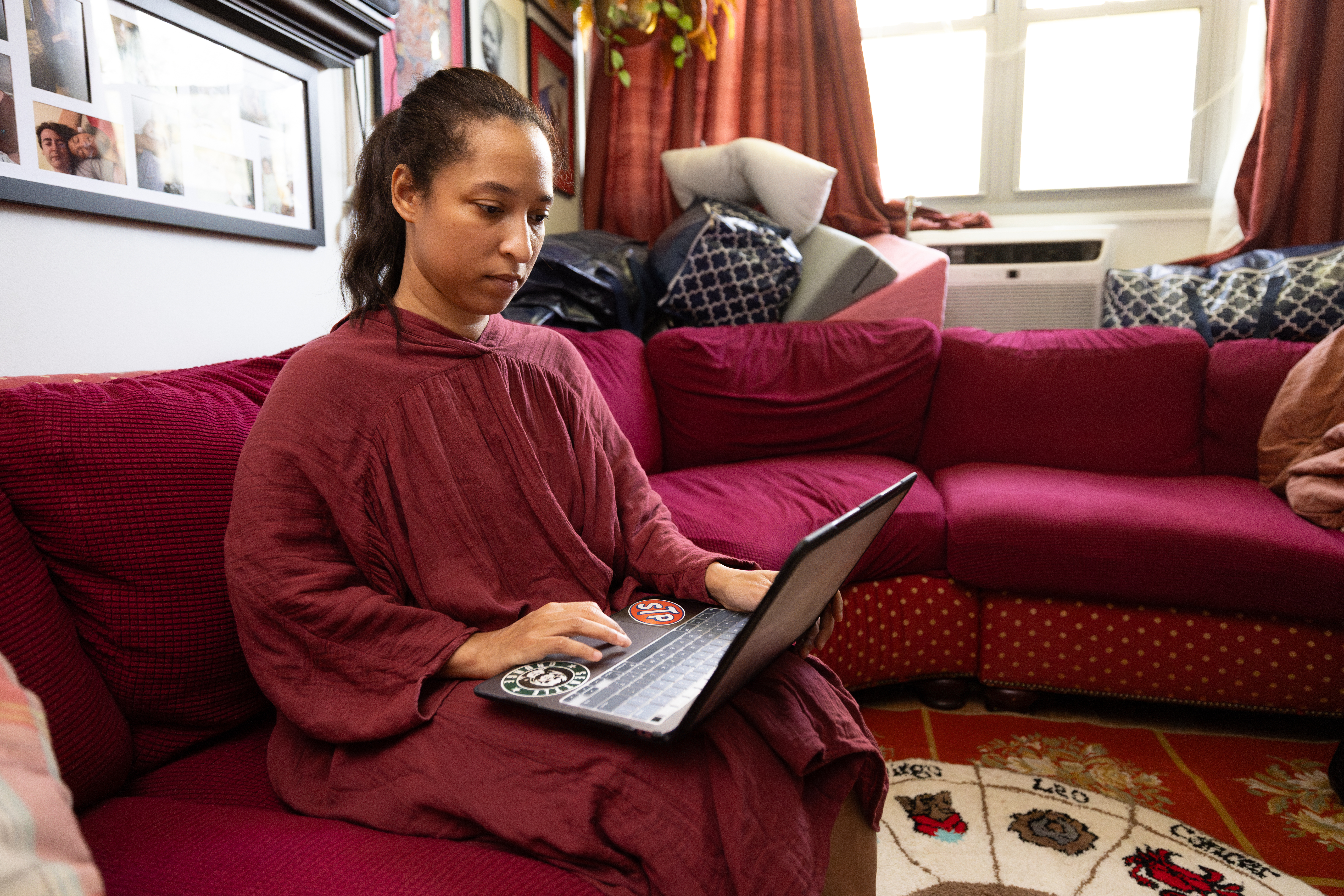 Woman looks down at laptop while sitting on her couch. Window is behind her. Stickers on her laptop.