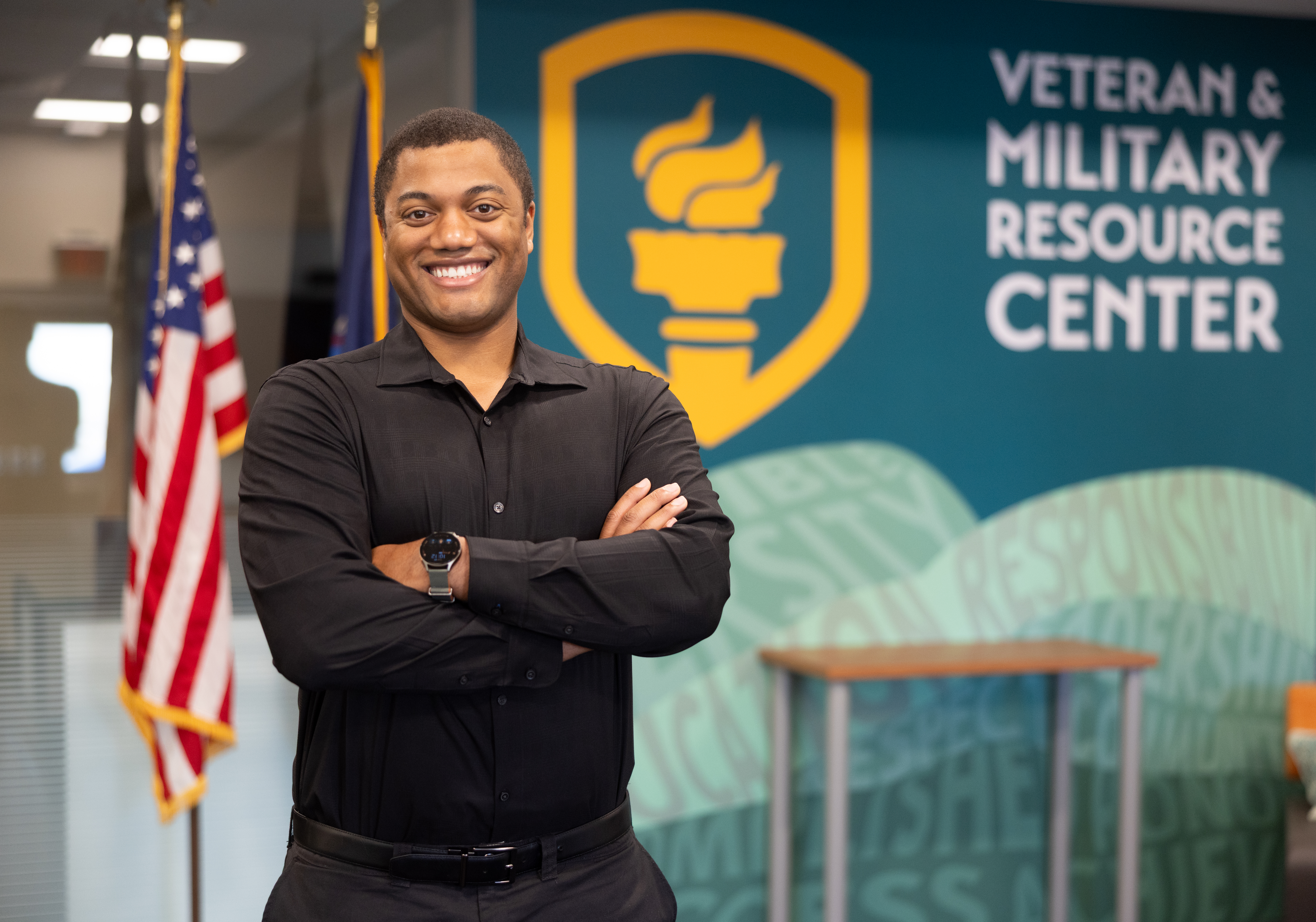 man stands smiling with his arms crossed with flags in the background. the wall futures a mural that has the SUNY Empire Shield and torch and "Vetern & Military resource center."