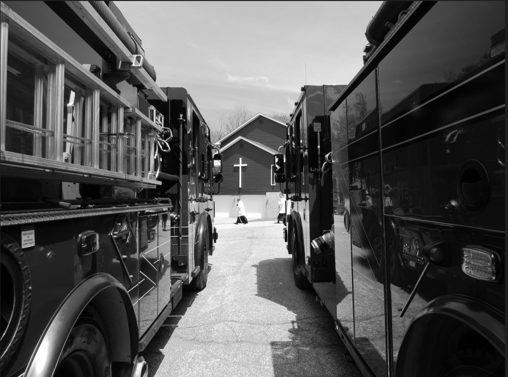 Picture looking down between two fire trucks parked next to each other. A building with a white cross on it is seen with a priest in a white robe walking from left to right underneath the cross.